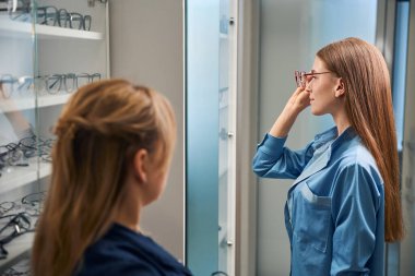 Genç bir kadın aynanın önünde gözlükleriyle görünüşünü incelerken, bir gözlükçü de izliyor.