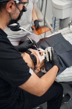 Koruyucu gözlüklü kadın, bir doktor ve bir asistan ağzına bir fotopolimer koyuyorlar.