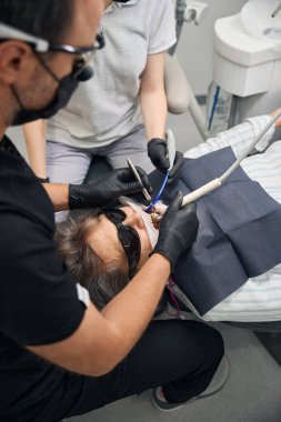 Yaşlı kadın dişlerini bir diş kliniğinde tedavi ettiriyor. Doktor ve asistanı koruyucu eldiven takıyor.