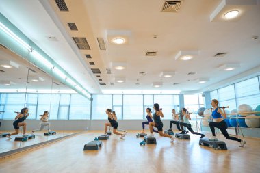 İnce kadınlar platformlarda jimnastik sopalarıyla ayna önünde antrenman yapıyorlar, rahat spor ayakkabıları giyiyorlar.