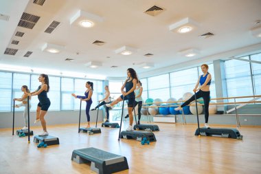 İnce kadınlar platformlarda jimnastik sopalarıyla bir takım egzersizler yapıyorlar, rahat spor ayakkabıları giyiyorlar.