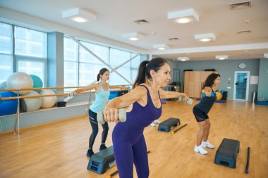 Eğitim odasında dambıllarla egzersiz yapan genç kadınlar, eğitim için de platformlar ve sopalar kullanılıyor.