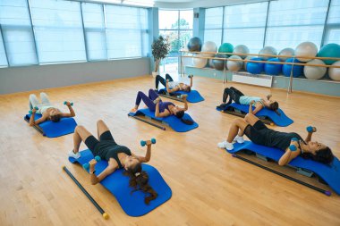 Bir grup kadın halterlerle güç egzersizleri yapıyor, özel platformlarda uzanan çeşitli spor aletleri derslerde kullanılıyor.