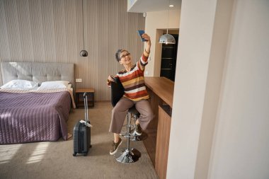 Otel odasında oturmuş akıllı telefondan selfie çekerken mutlu yetişkin bir kadın. Seyahat kavramı