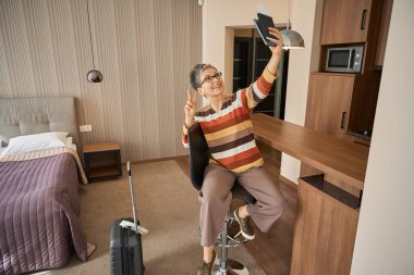 Gülümseyen yetişkin kadın, zafer jesti yapıyor. Otel odasında selfie çekiyor. Seyahat kavramı