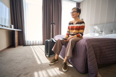 Otel odasındaki yatakta elinde telefon ve biletlerle oturan gözlüklü yaşlı bir kadın. Seyahat kavramı