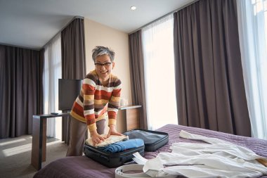 Otel odasında kıyafetlerini toplayıp kameraya bakan mutlu yaşlı bayan. Seyahat kavramı
