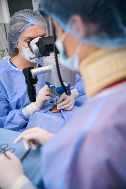 Cerrah, bir cerrahi mikroskop kullanarak, yanında deneyimli bir asistanla bir operasyon gerçekleştirir.