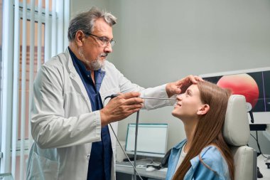 Beyaz önlüklü Otolaryngologist, özel bir tıbbi araç kullanarak oturan bir hastaya göz muayenesi yapar.