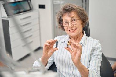 Yaşlı bir kadın diş implantı bileşenlerinin bir modelini elinde tutuyor, dişçilikte modern teknolojilerin kullanımı
