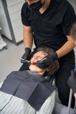 Özel bir koltukta dişçi muayenehanesindeki yaşlı kadın, doktor ve asistanı dişlerini tedavi ediyorlar.