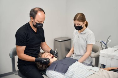 Uzman, yaşlı bir bayanın dişlerini tedavi ediyor, genç bir asistan da ona işinde yardım ediyor.