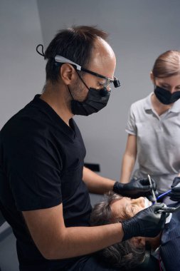 Dişçi ve asistan dişlerini yerine takıyor, erkek ise dürbün kullanıyor.