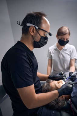 Diş hekimi ve asistanı kadının dişlerini yerine takıyor, erkek ise dürbünlü diş gözlüğü kullanıyor.