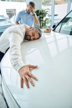 Otomobil galerisinde beyaz arabanın kaputunda yatan mutlu genç adam. Araba satışı, iş konsepti