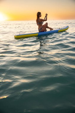 Genç beyaz kadın, sakin bir günbatımında okyanusta kürek çekmeyi seviyor. Sakin sular, gökyüzünün sıcak tonlarını yansıtarak, barışçıl bir atmosfer yaratır..