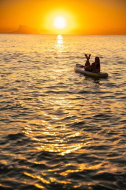 Genç kadın sakin okyanus sularında dinlenirken kürek çekmenin tadını çıkarıyor. Altın gün doğumu huzurlu bir atmosfer yaratır, huzurlu havayı arttırır..
