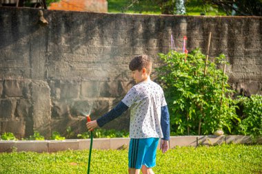 Brezilyalı çocuk güneşli bir öğleden sonra arka bahçesinde su hortumuyla oynuyor. Beyaz, beyaz, latin çocuk suyla oynuyor. Eğlenceliydi. Mutlu bir çocukluk. Arka bahçede oyun günü..