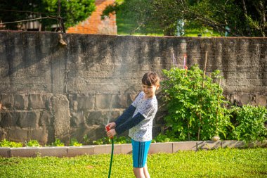 Brezilyalı çocuk güneşli bir öğleden sonra arka bahçesinde su hortumuyla oynuyor. Beyaz, beyaz, latin çocuk suyla oynuyor. Eğlenceliydi. Mutlu bir çocukluk. Arka bahçede oyun günü..