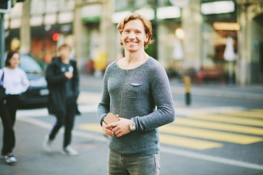 Outdoor portrait of happy handsome red-haired man wearing blue pullover, holding phone, city street and crosswalk on background