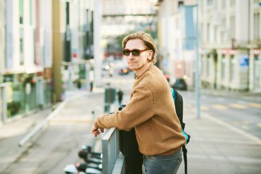 Outdoor portrait of handsome young man wearing sunglasses and beige fuzzy fleece sweater, posing on city street background
