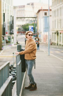 Outdoor portrait of handsome young man wearing sunglasses and beige fuzzy fleece sweater, posing on city street background