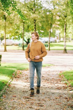 Outdoor portrait of handsome young man walking outside through public park, wearing brown pullover and backpack