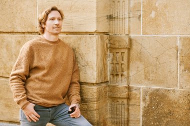 Outdoor fashion portrait of handsome red-haired man posing next to brown wall, wearing beige fuzzy fleece sweater