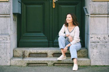 Outdoor portrait of beautiful 40 year old woman sitting on step next to green door, old money fashion style