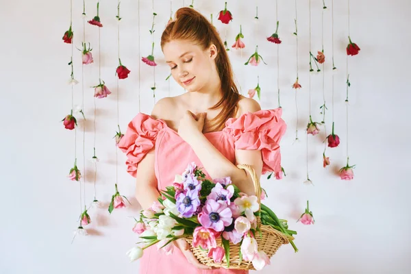 15-16 yaşlarında, pembe mercan elbiseli, beyaz arka planda asılı güllerle poz veren, bahar çiçekleri, güzellik ve moda kavramlarıyla dolu sepetli güzel bir genç kızın stüdyo portresi.