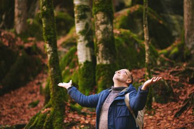 Yakışıklı, orta yaşlı, ormanda yürüyüş yapan, ceketli, sırt çantalı ve gözlüklü bir adam.