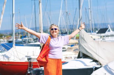 50-55 yaşlarında, renkli kıyafetler giyen, göl veya deniz limanında poz veren, seyahat ve tatil hayatı tarzında orta yaşlı bir kadının açık hava moda portresi.
