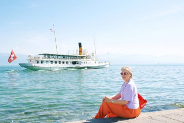 50-55 yaşlarındaki olgun bir kadının açık hava portresi gölün kenarında oturmuş İsviçre 'nin Cenevre gölünde yüzen bir Fransız teknesini seyrediyor.