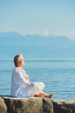 Açık havada, gölün ya da denizin kenarında meditasyon yapan, beyaz kıyafetler giyen, ruhanilik, uyanış ve yaşam dengesi olan mutlu olgun bir kadının portresi.