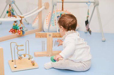 Adorable baby playing with different kind of toys in daycare or kindergarten, back view clipart