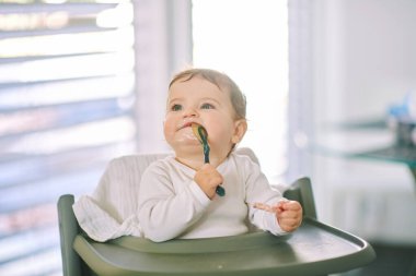 Sevimli, dağınık bir bebeğin portresi yüksek sandalyede oturuyor, kaşık tutuyor.