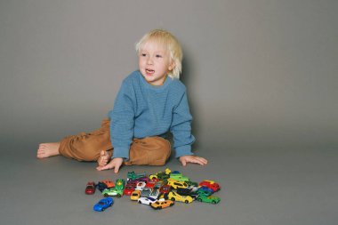 Bir sürü oyuncak arabayla oynayan sevimli küçük çocuğun gri arka planında stüdyo portresi