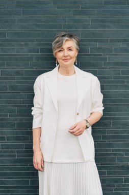 55-60 yaşlarında beyaz ceketli, gri arka planda poz veren gri saçlı bir kadın.