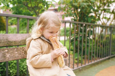 Sweet portrait of adorable little toddler girl eating baguette chease sandwich, sitting on bench in outdoor park