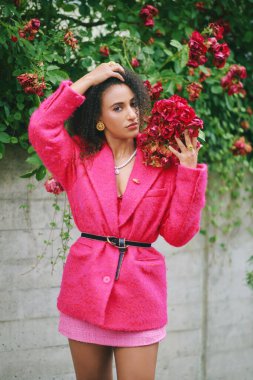 Parlak pembe ceket giyen, kırmızı güllerle poz veren güzel Afrikalı bir kadının açık hava moda portresi.
