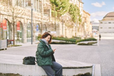 Açık havada cep telefonu kullanan, bankta oturan, sıcak yeşil ceket giyen mutlu bir kadının portresi.
