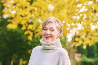 Mutlu 55-60 yaşındaki bir kadının sonbahar parkında aktif ve sağlıklı bir şekilde poz verdiği mutlu bir portre.