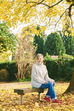 55-60 yaşlarında, sonbahar parkında dinlenen, bankta oturan bir kadının açık hava portresi