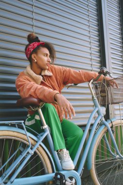 Bisiklette oturan, duvara yaslanan güzel Afrikalı bir kadının açık hava moda portresi.
