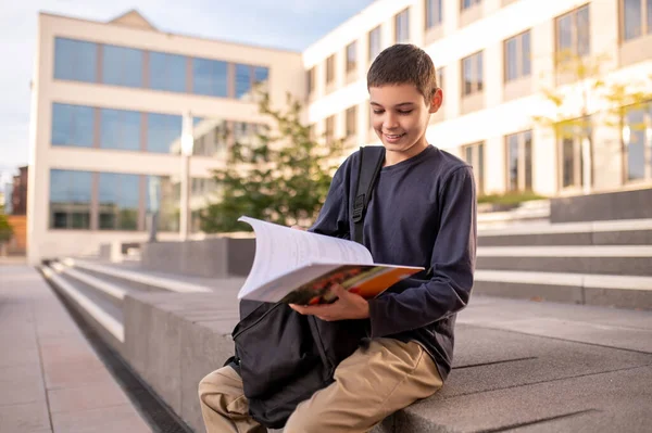 Uśmiechnięty Nastolatek Siedzący Werandzie Budynku Przedzierający Się Przez Książkę — Zdjęcie stockowe