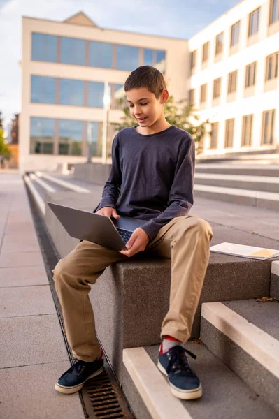 Focused adolescent boy sitting on the porch of a building and typing on the laptop