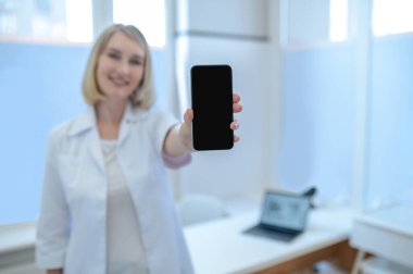 Beyaz laboratuvar önlüğü giymiş, cep telefonunu kameranın önünde tutan memnun bir kadın doktor.