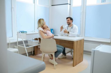 Gülümseyen dost canlısı pediatrik doktor, muayene sırasında çocukla birlikte bir kadına beslenme takviyesi yapıyor.