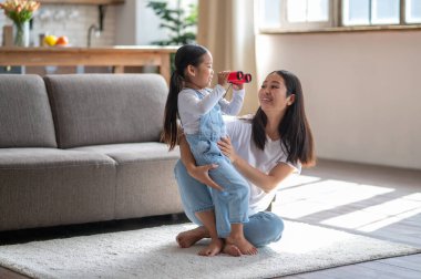 Sevecen bir anne halının üzerinde oturuyor ve elinde dürbünle kızını kucaklıyor.