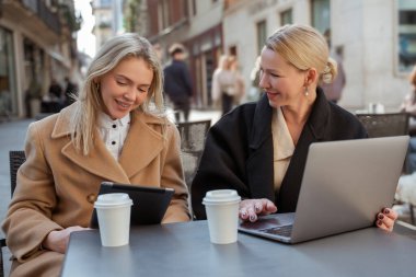 Dışarıda buluşacağız. İki sarışın iyi giyimli kadın bir kafede oturuyor.